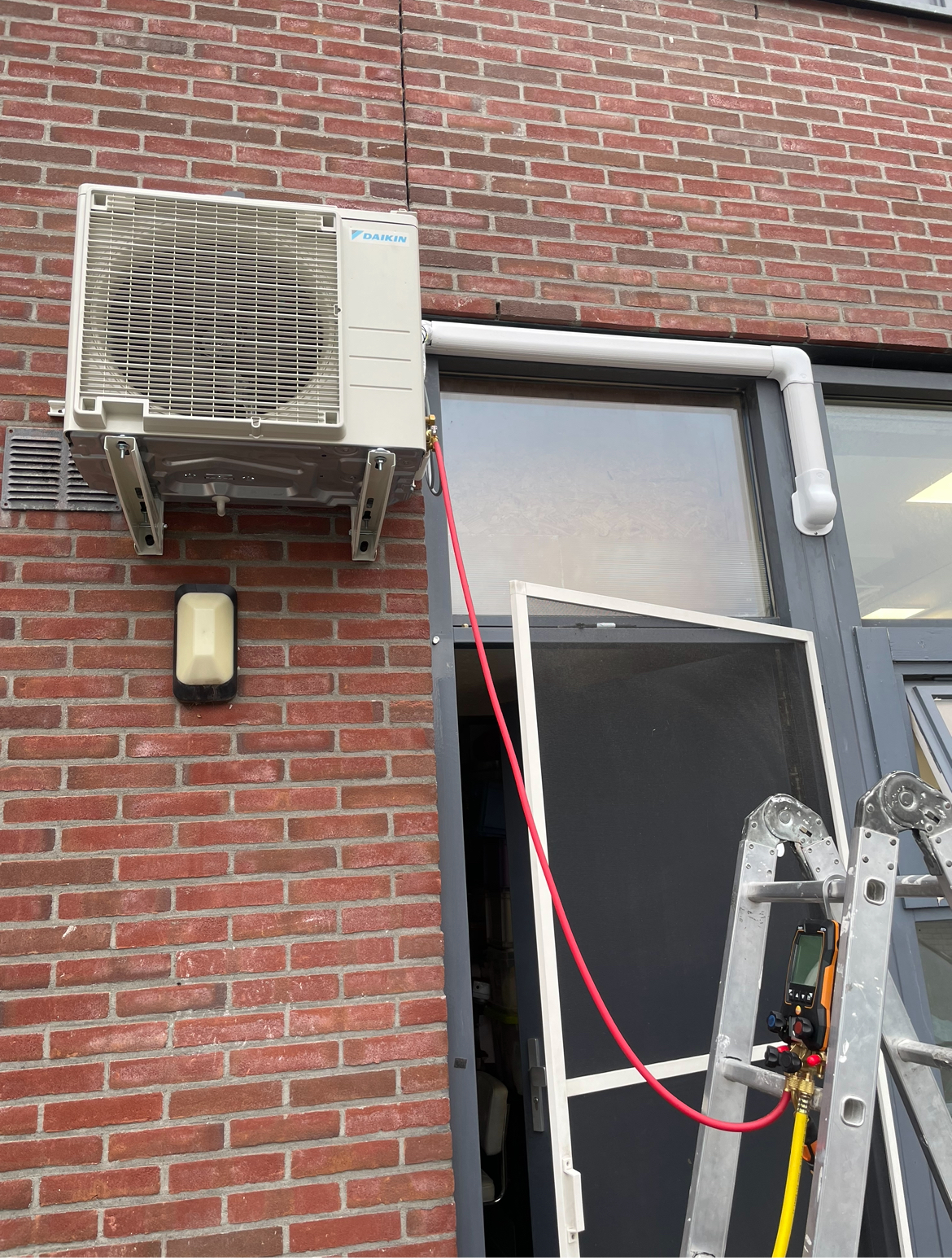 Daikin-buitendeel op beugels aan een rode bakstenen gevel, met ladder en slangen